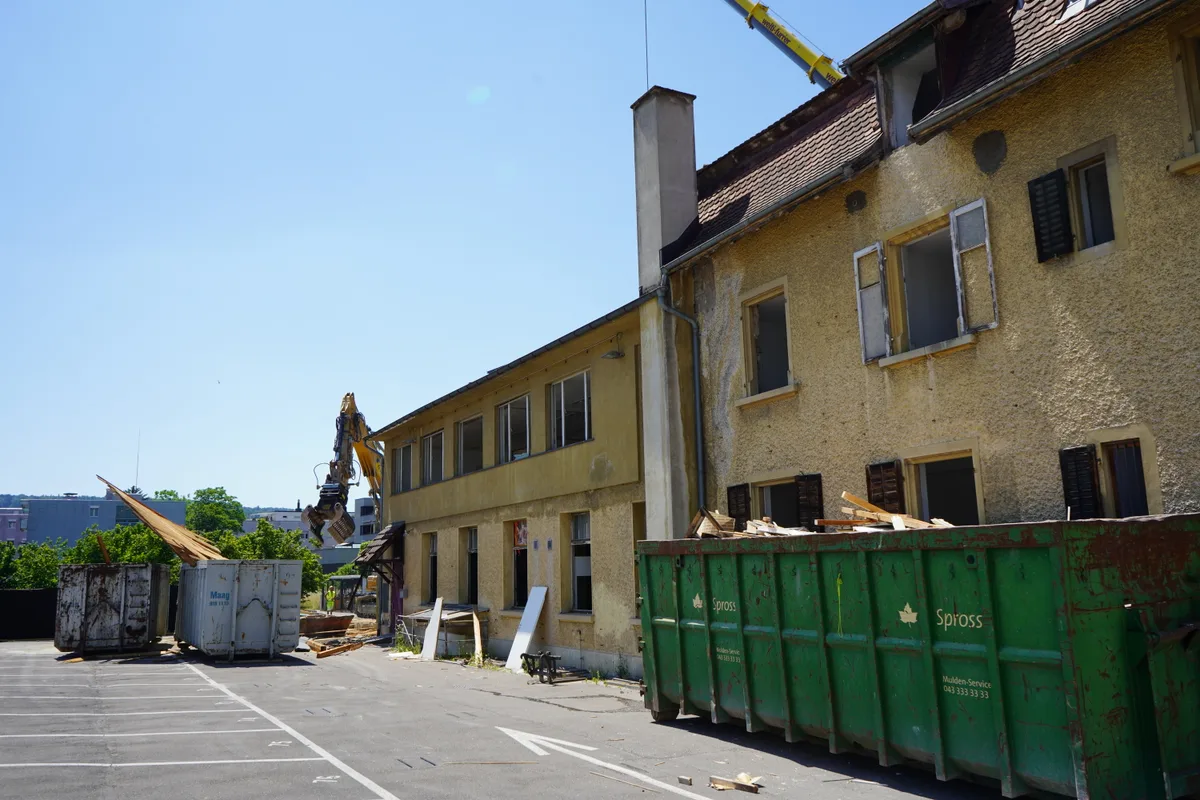 Baustelle mit Abbruchhaus, Bagger und Schuttmulde.